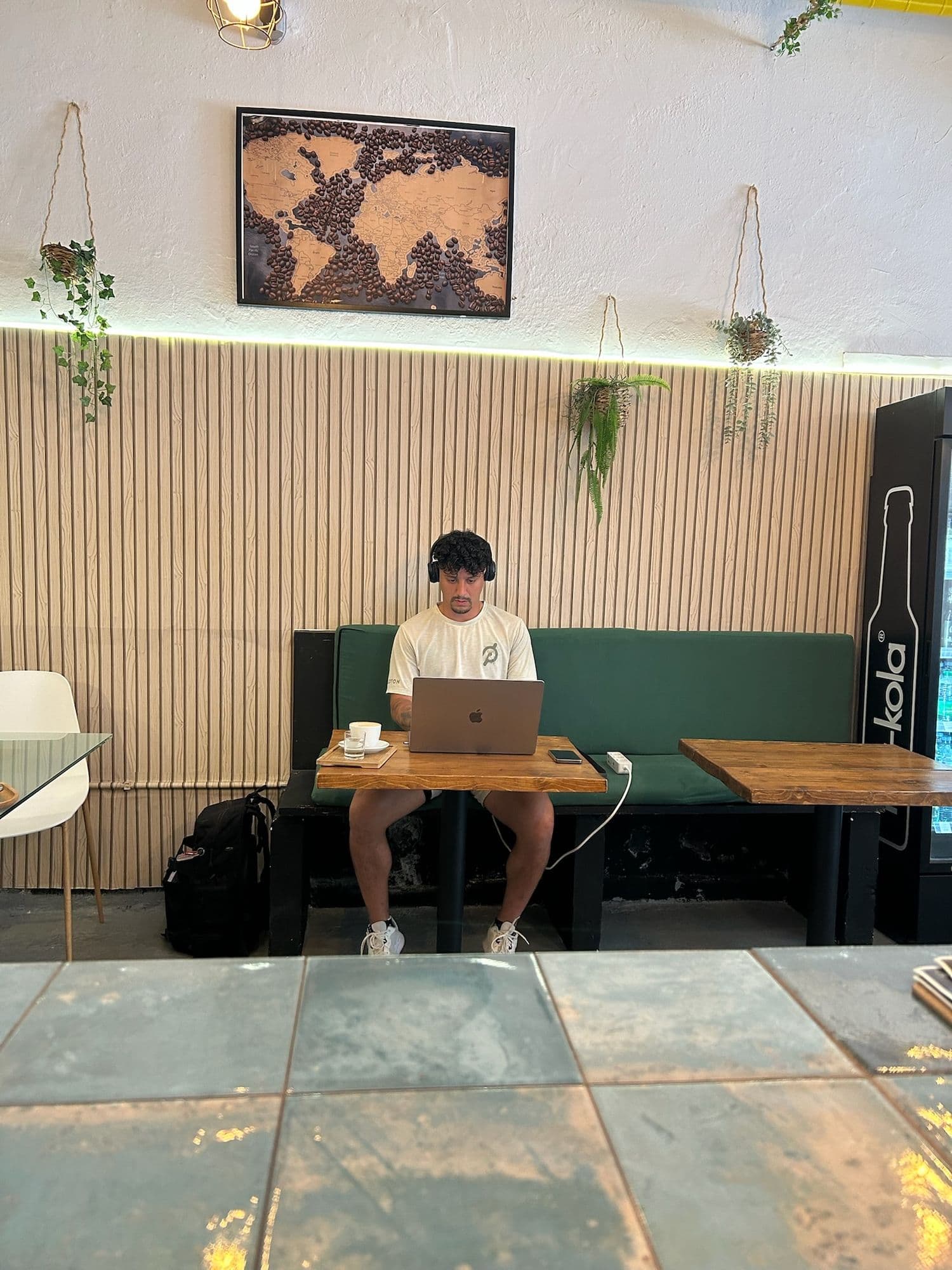Un chico con auriculares trabaja con su portátil en el banco verde, con un café en la mesa.