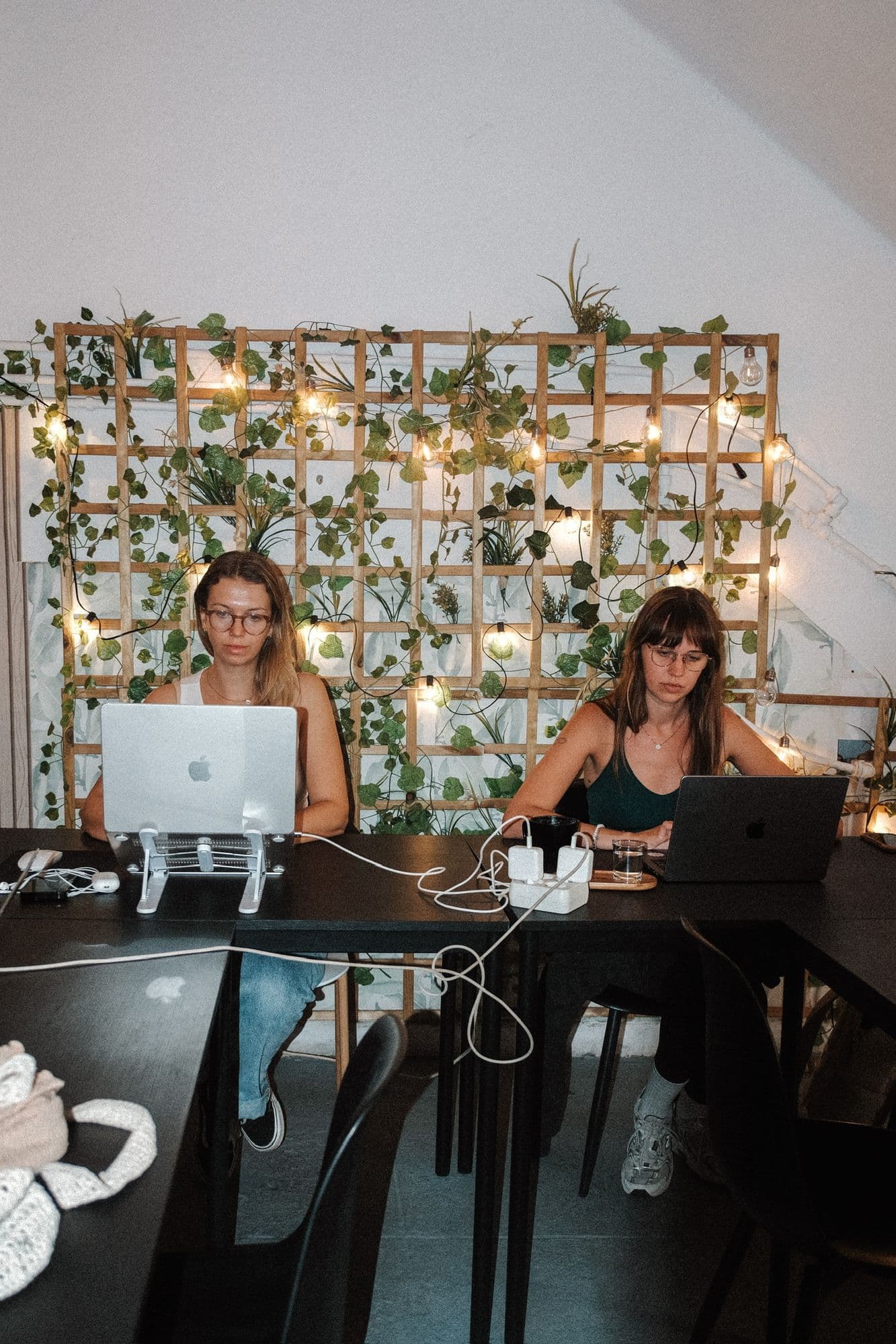 Dos chicas trabajan con sus portátiles en la mesa larga, delante de la pared de enredaderas y bombillas.