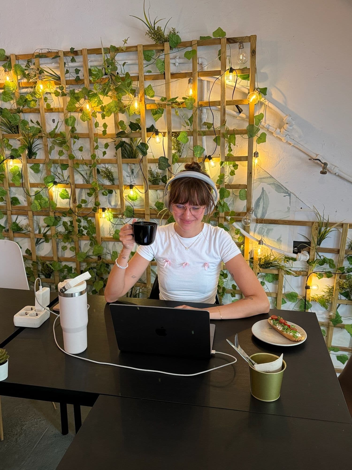 Una chica con auriculares sonríe con una taza en la mano, junto a su portátil y una tostada, delante de la pared de enredaderas y bombillas.