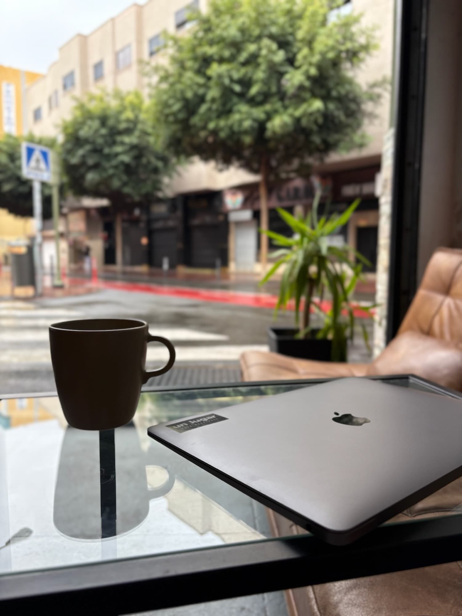 Una taza de café y un portátil junto a la ventana, con la calle al otro lado del cristal.