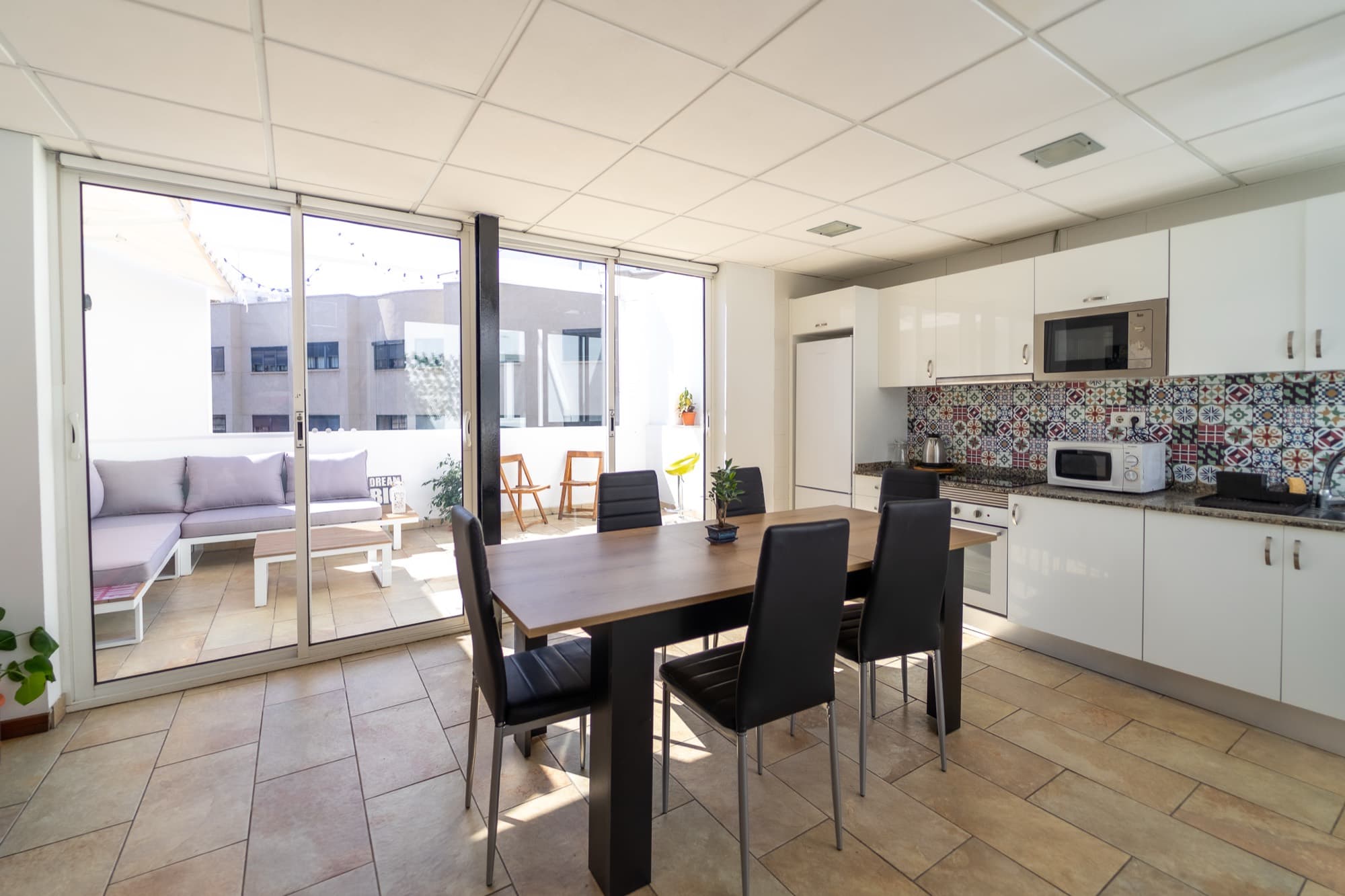 Cocina compartida con muebles blancos, azulejos de colores y una mesa de comedor junto a las puertas de la terraza.