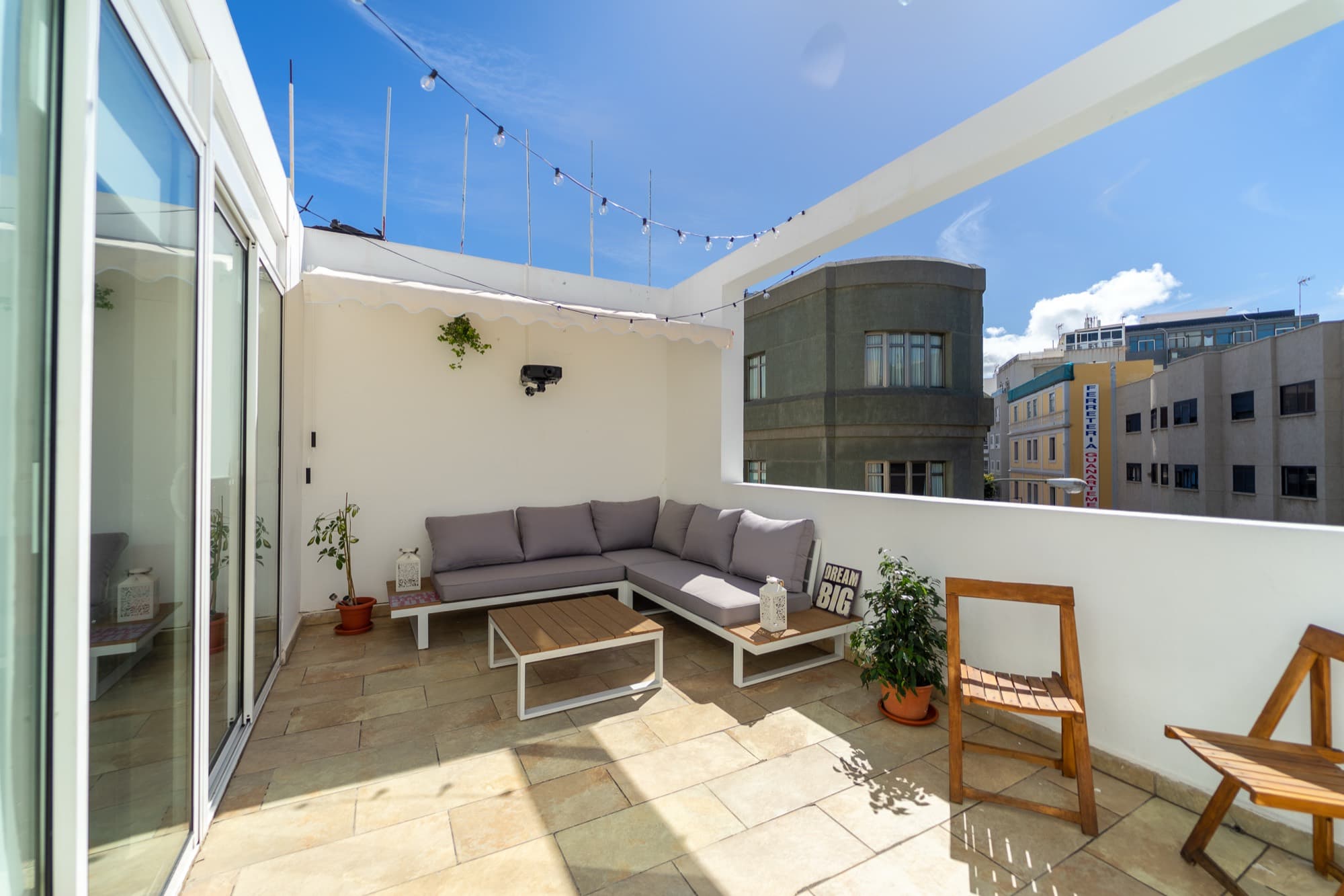 Die Terrasse von House Canteras in der Sonne: graues Outdoor-Sofa, Holzklappstühle und Lichterketten unter blauem Himmel.