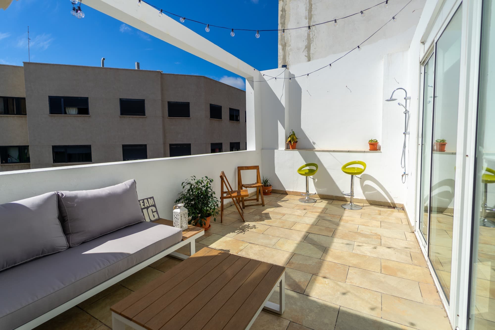 Un rincón de la terraza con sofá gris, mesa baja, taburetes altos y ducha exterior.