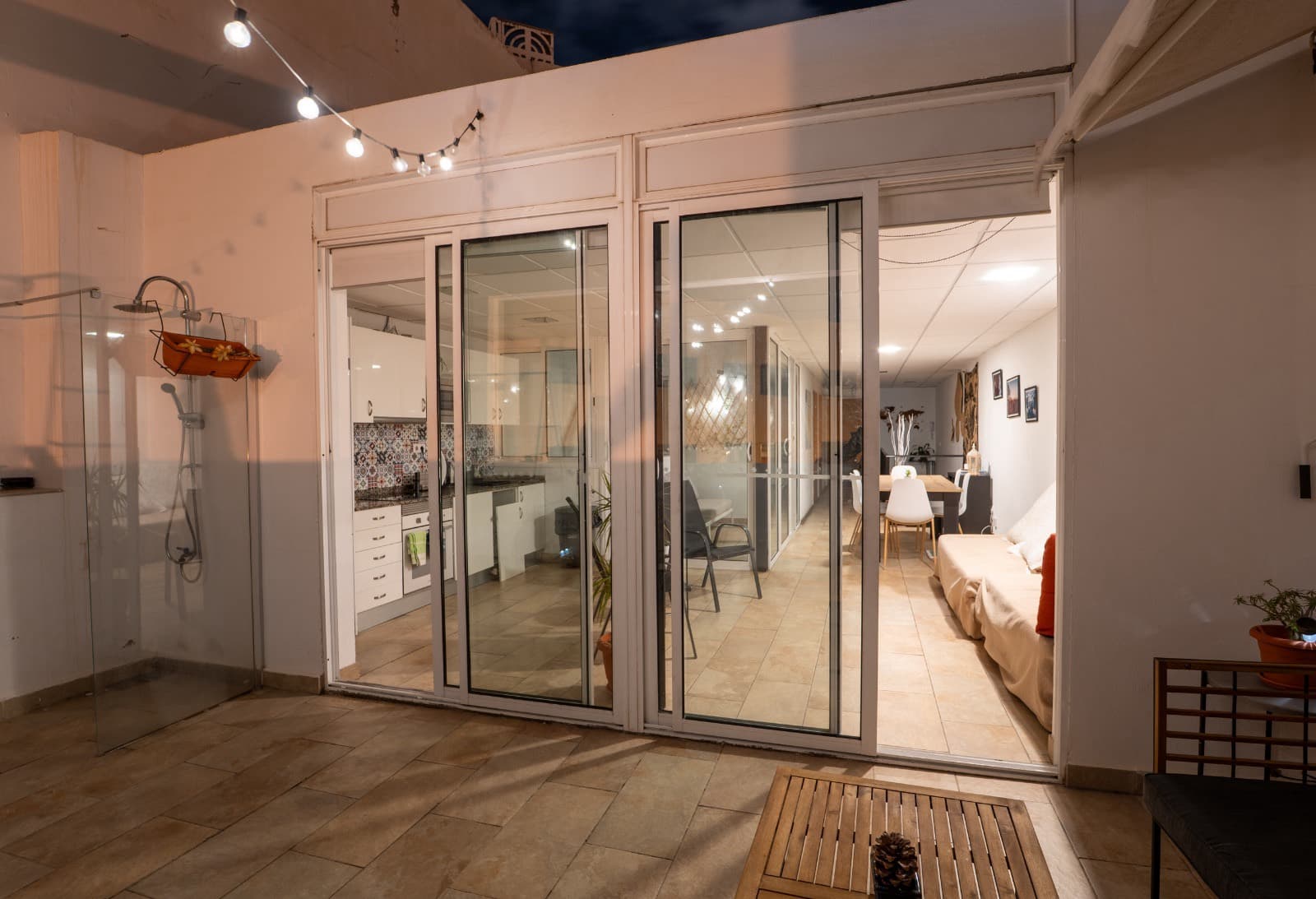 La terraza de noche, iluminada con guirnaldas, con la cocina y el salón encendidos al otro lado de las puertas de cristal.