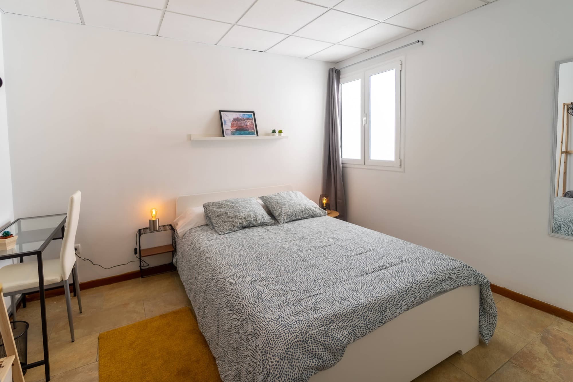 Habitación Doppio: cama doble, escritorio de cristal con silla blanca y ventana con cortina gris.